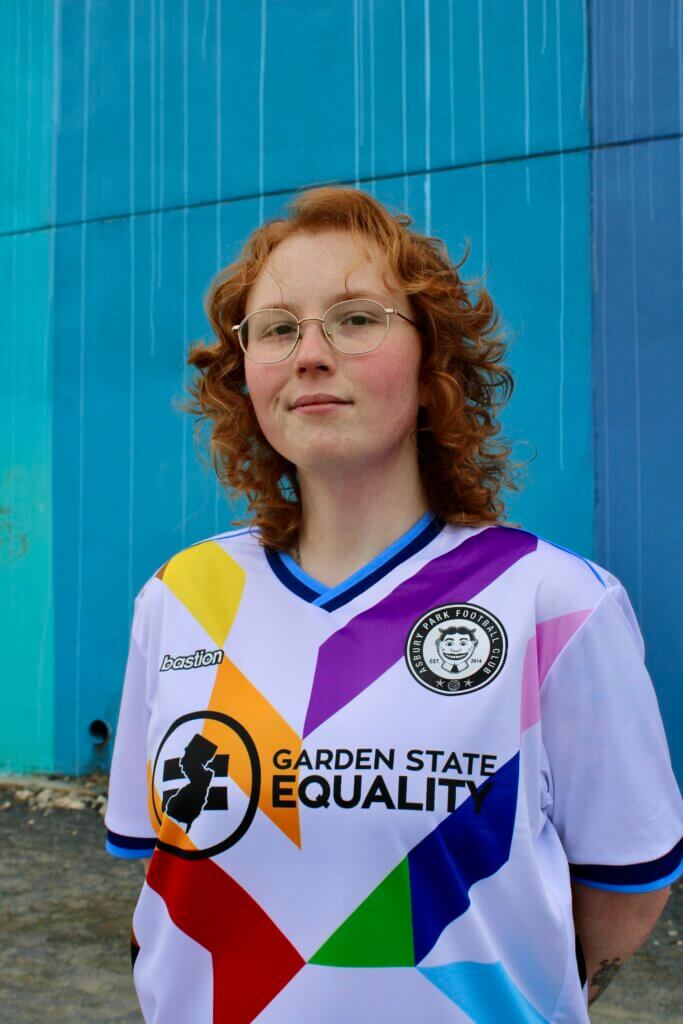 aedy miller, a white redheaded trans woman with glasses, smiles at the camera. She is wearing a Garden State Equality soccer jersey. In the background is a wall painted multiple shades of blue.