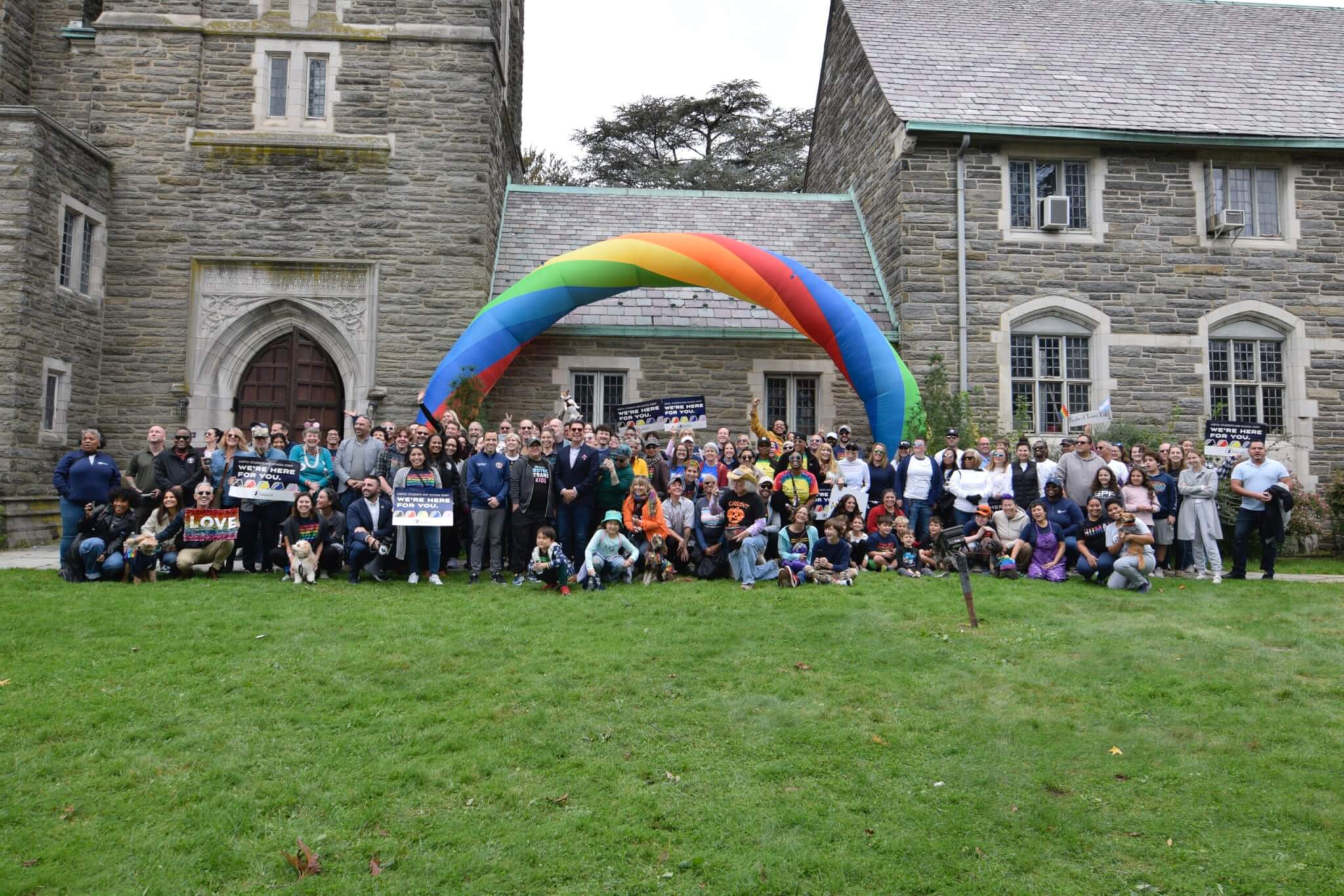 A large group of LGBTQ+ people, allies, families, and other community members pose for a group photo underneath an inflatable rainbow arch before the Equality Walk in Montclair, New Jersey. Behind them is the historic First Congregational Church of Montclair.