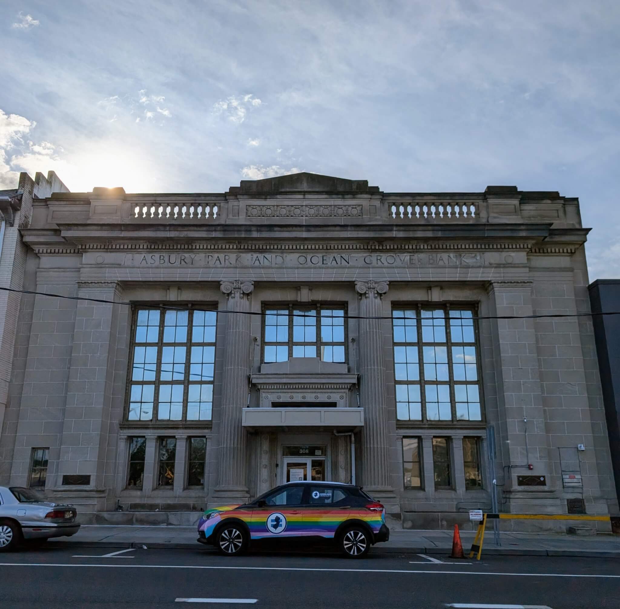 In front of the historic Asbury Park and Ocean Grove Bank building in Asbury Park sits the Garden State Equality rainbow car. The sun rises behind the building.